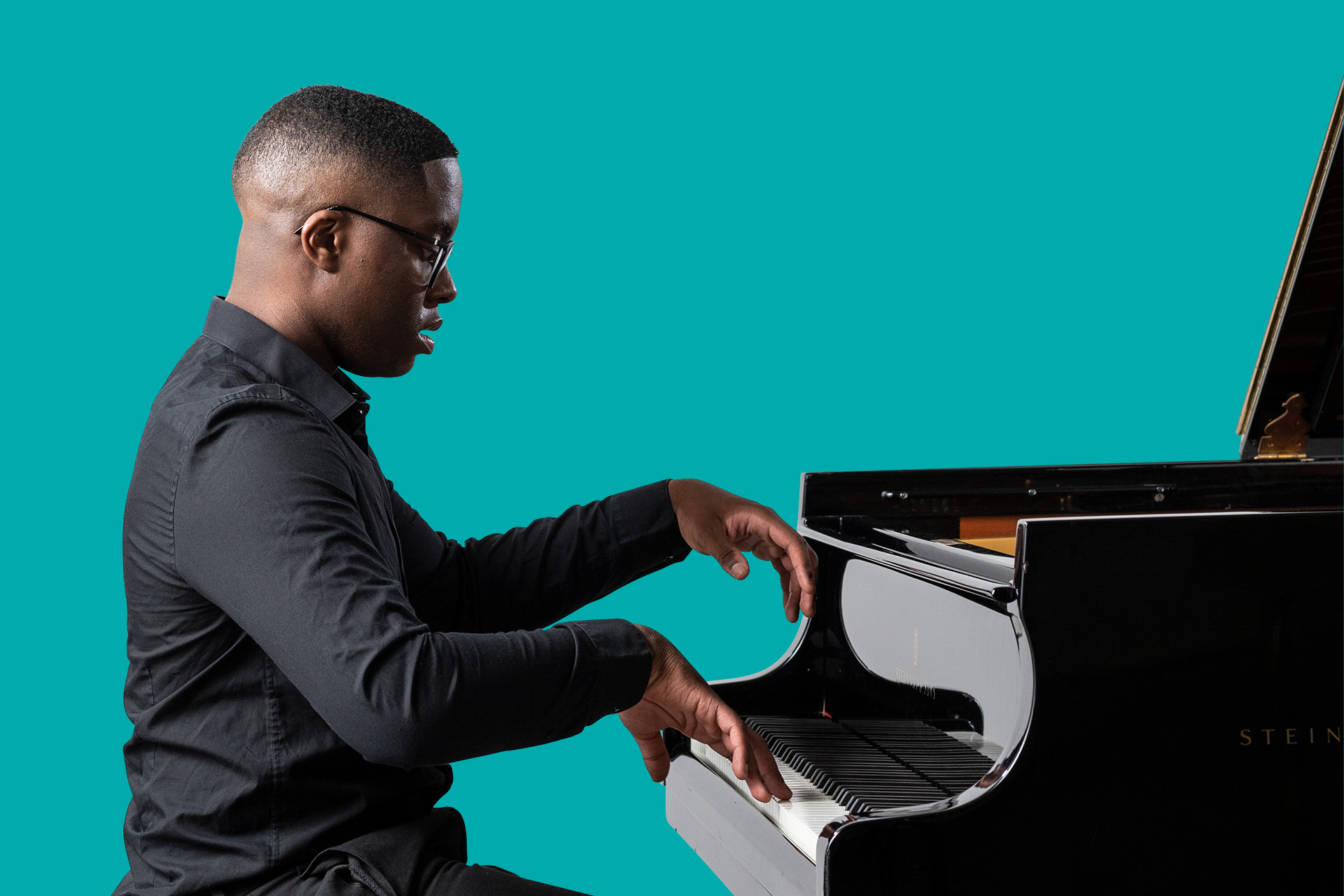 A male students wearing a black shirt playing the piano against a teal background
